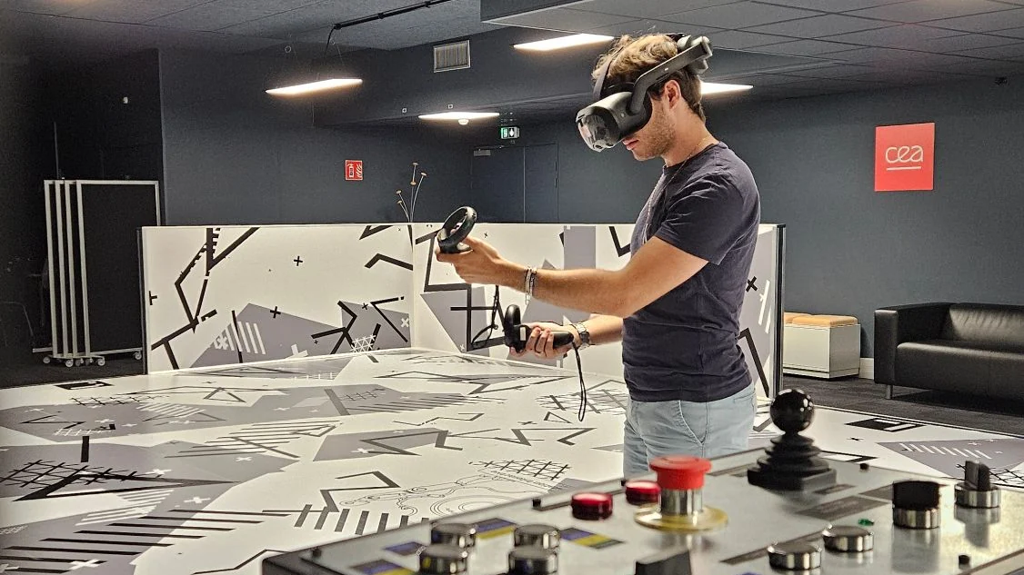 Thibaut, casque de réalité virtuelle sur la tête et manettes en main, dans la salle de réalité virtuelle du laboratoire&nbsp;; au mur, le logo du CEA.