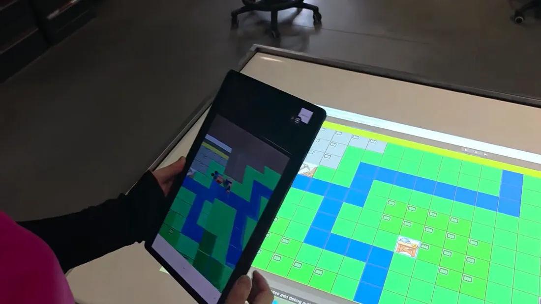 Une tablette tenue au-dessus de la carte projetée sur une table&nbsp;: son écran montre la même carte en réalité augmentée, avec des bâtiments en 3D.